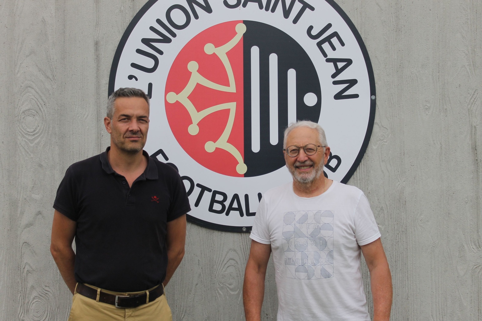 Deux hommes debout souriants posent devant le logo du club de football Union Saint Jean, appos&eacute; sur un mur en bois clair.