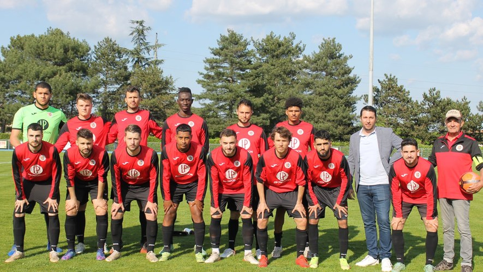 Photo d&rsquo;&eacute;quipe de football en maillot rouge et noir posant sur le terrain, entour&eacute;e de membres du staff, sous un ciel d&eacute;gag&eacute; avec des arbres en arri&egrave;re-plan.